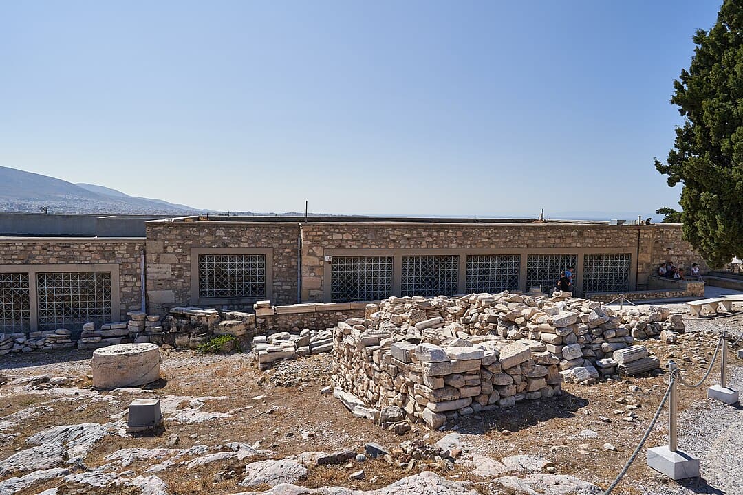 Ancient stone ruins with modern preservation building and visitors at archaeological site under blue sky inOld Acropolis Museum Greece