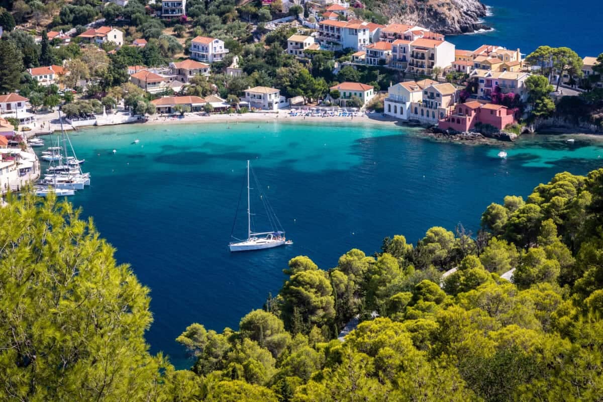 Assos Kefalonia Greece coastal village with sea, boats, white houses, and lush greenery.