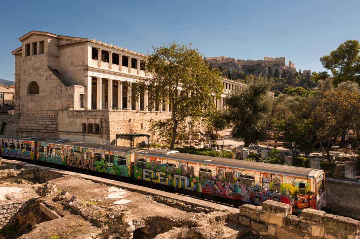 Graffiti-covered Athens subway train near Acropolis and ancient ruins showing modern and historic contrast.