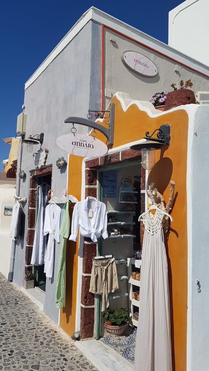 Spilia Cave Oia Santorini Greek boutique with pastel clothing display on cobblestone street under bright Mediterranean sunlight.