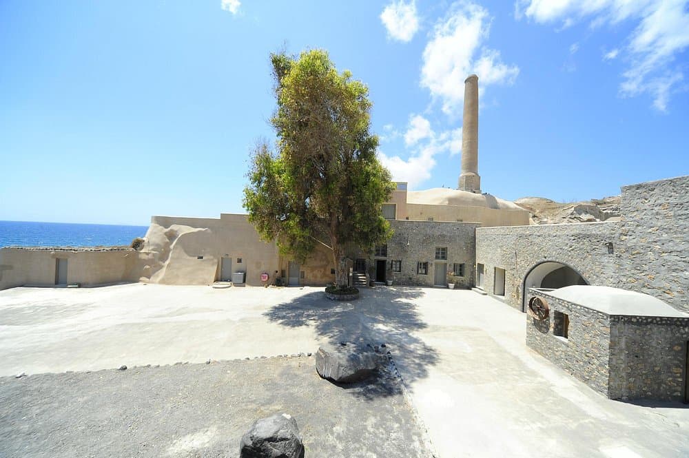 Historic coastal Tomato Industrial Museum, D.Nomikos courtyard with stone buildings, smokestack, large tree, and ocean view.