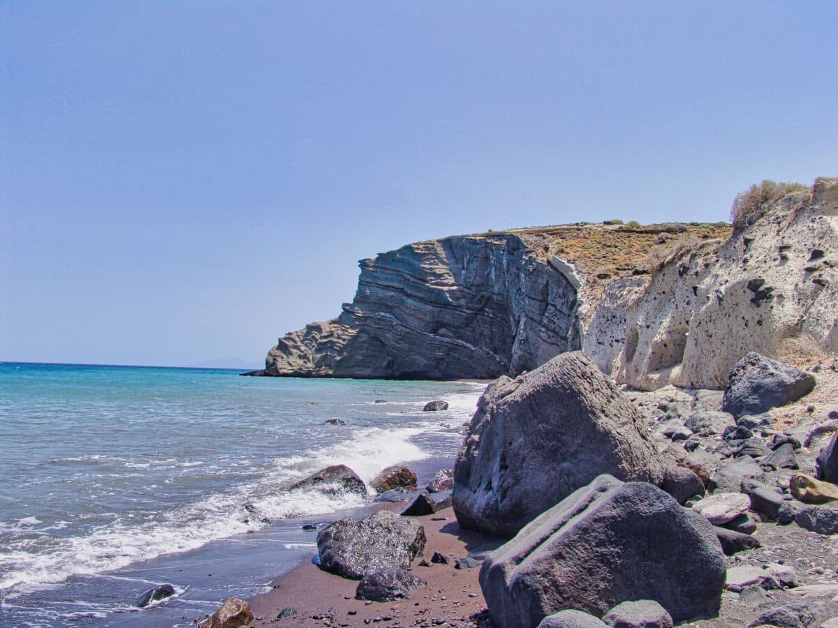 Koloumbos Beach in Santorini features rocky cliffs, dark sands, and crystal-clear Aegean waters.