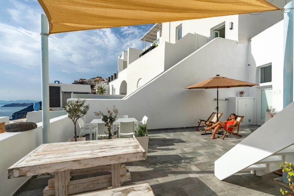 Akron Cliff Suite Mediterranean coastal terrace with sea view, relaxing woman, outdoor dining, whitewashed architecture, sunny weather.