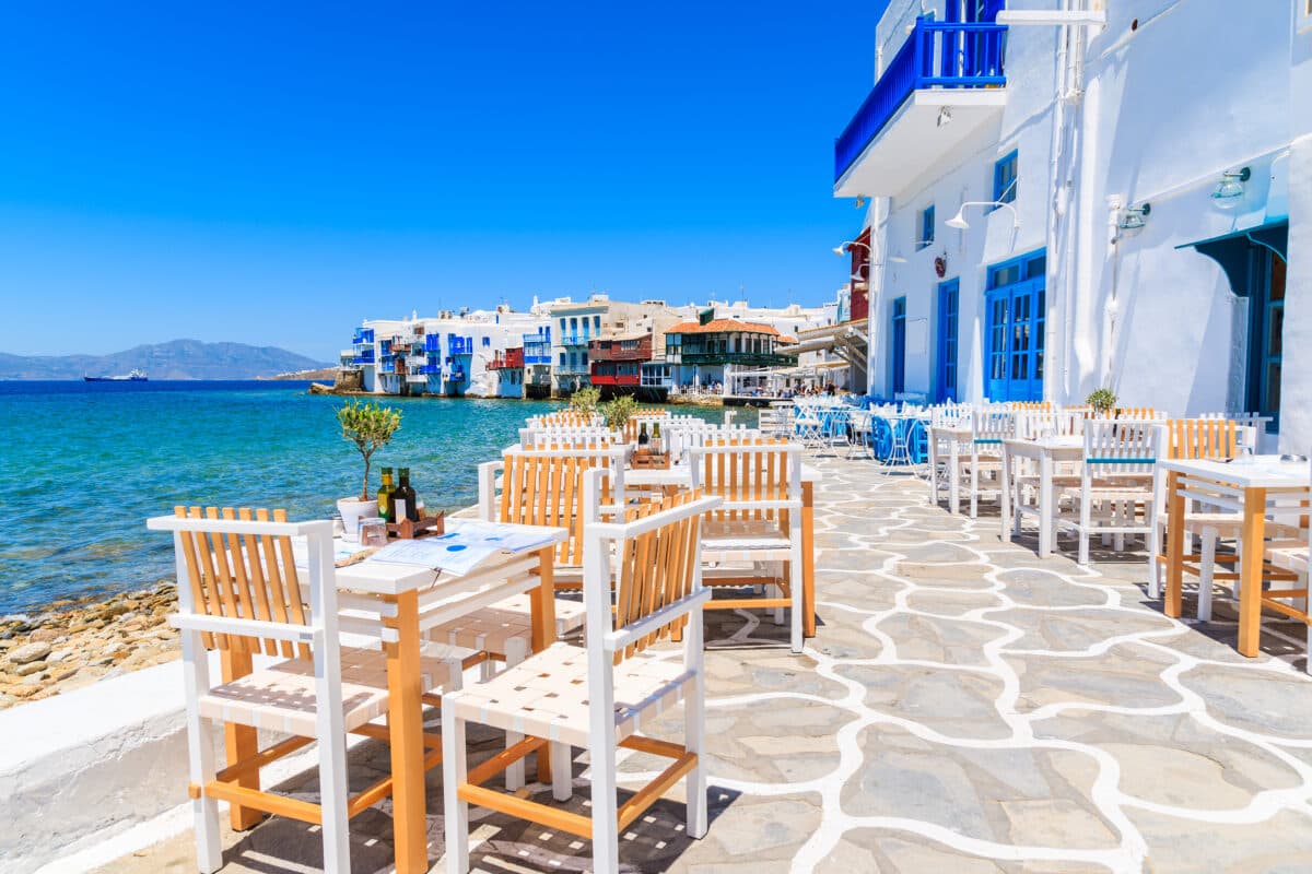 Greek seaside tavern in Little Venice, Mykonos, with white tables, blue shutters, and Aegean Sea.