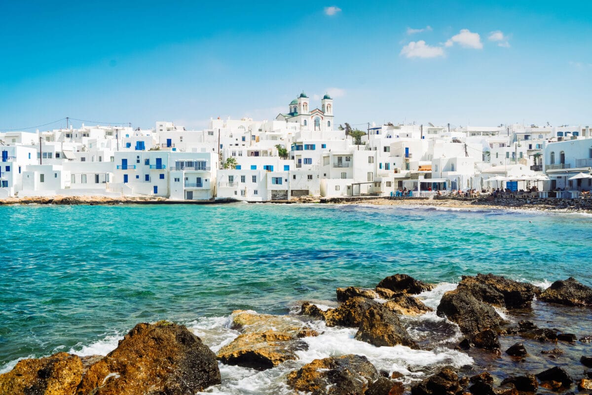Naoussa waterfront in Paros, Greece, features white buildings, blue shutters, a church, and turquoise sea.