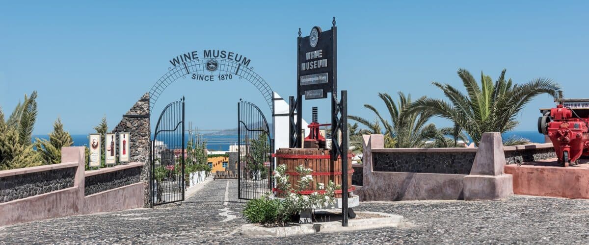 Koutsogiannopoulos Wine Museum & Winery entrance with rustic iron gate, vintage wine press, and coastal scenery.