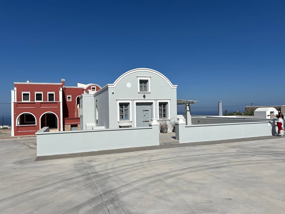 Canava Santorini Distillery white and red buildings with courtyard and person walking in sunny outdoor setting.